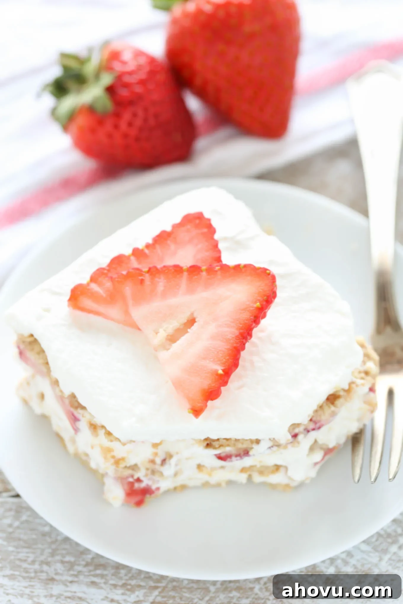 A slice of strawberry graham cracker icebox cake on a white plate with a fork. Two fresh berries rest in the background on a towel. 