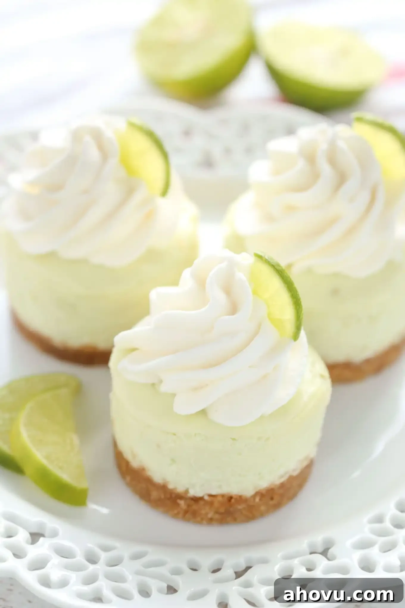 Three mini key lime cheesecakes on a white plate. Two lime halves are in the background, suggesting fresh ingredients and a zesty flavor.