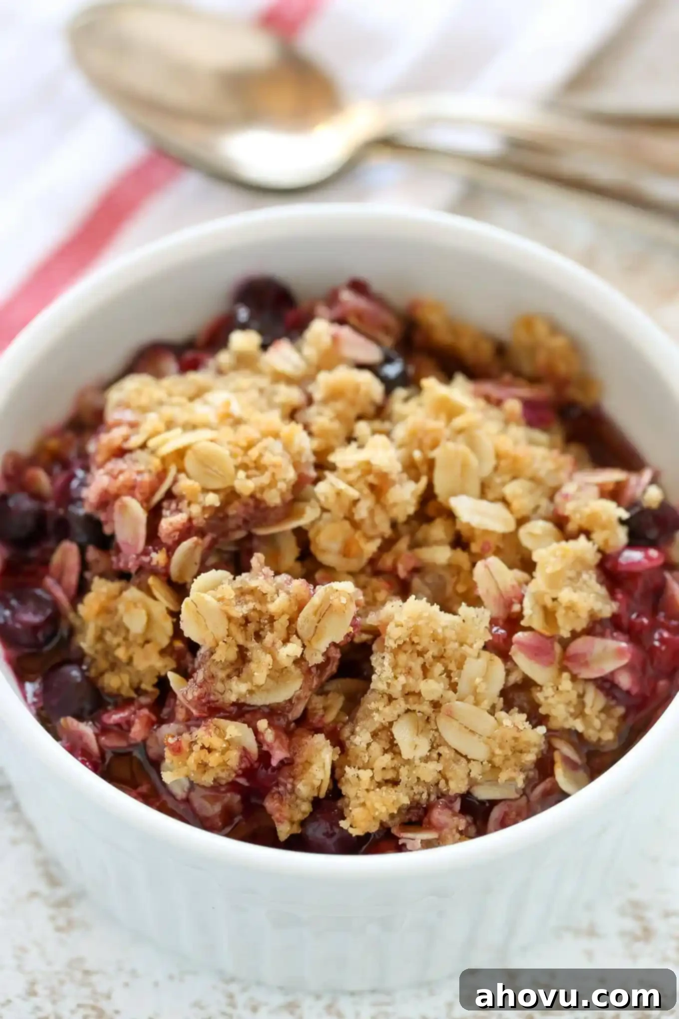 An individual serving of frozen berry crisp in a white ramekin. Two spoons and a towel rest in the background. 