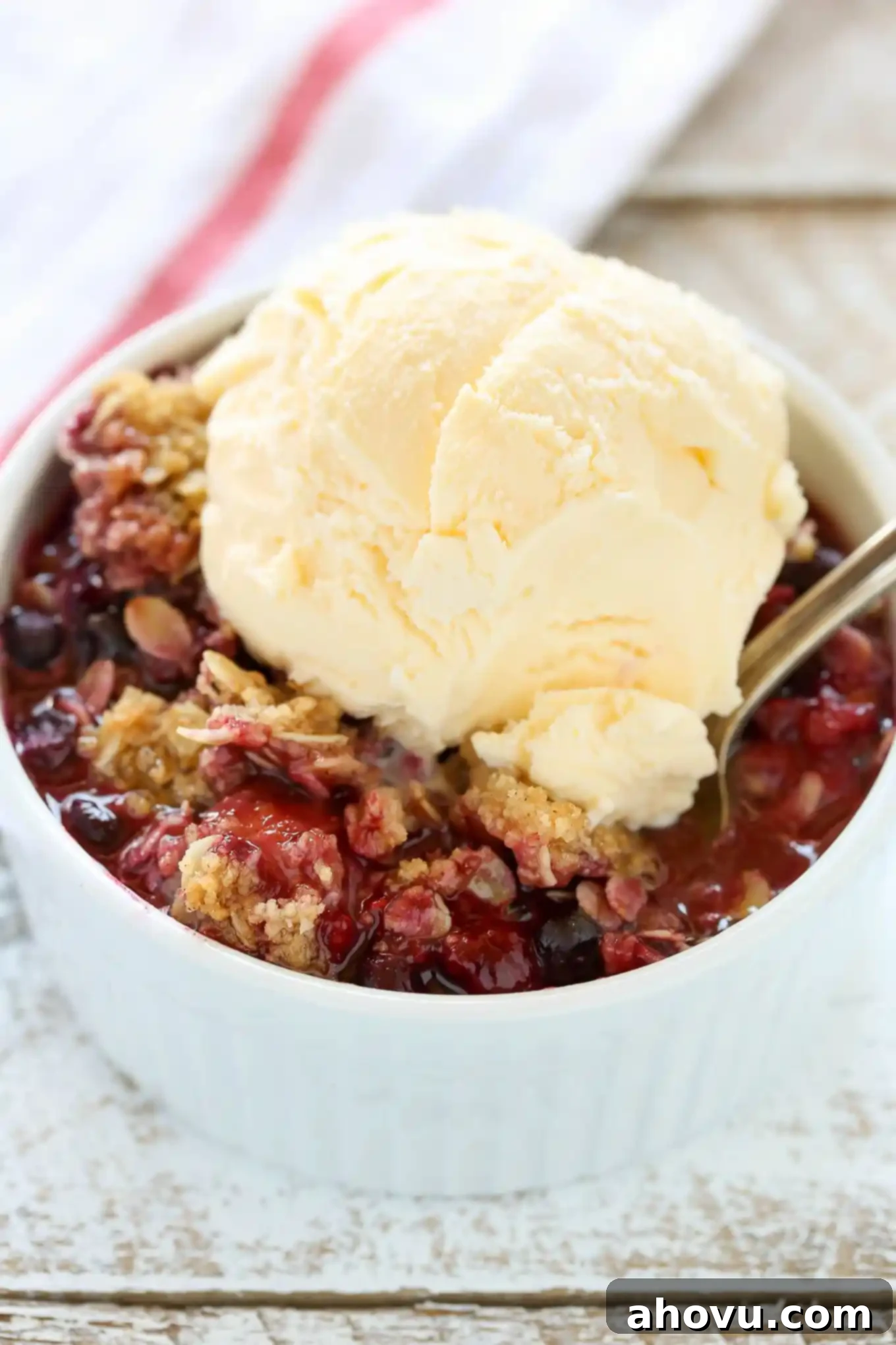 A white ramekin filled with mixed berry crisp with frozen berries, topped with a scoop of vanilla ice cream. 