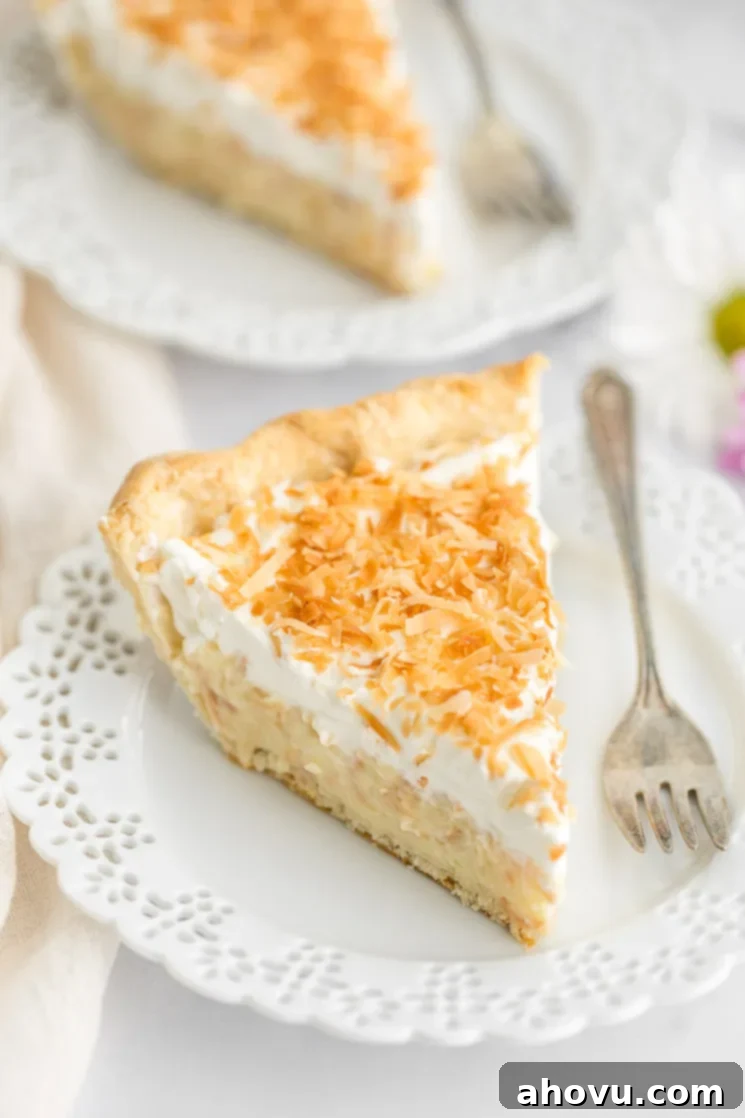 A generous slice of homemade coconut cream pie, showcasing its layers of creamy filling and whipped topping, served on a decorative white plate. Another pie slice is visible in the soft-focus background.
