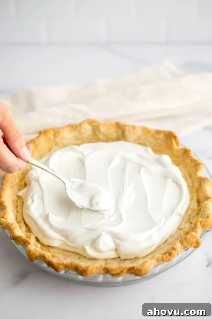 A close-up shot of a fluffy, white cloud of whipped cream being generously swirled onto the top of a coconut cream pie, creating a beautiful peak.