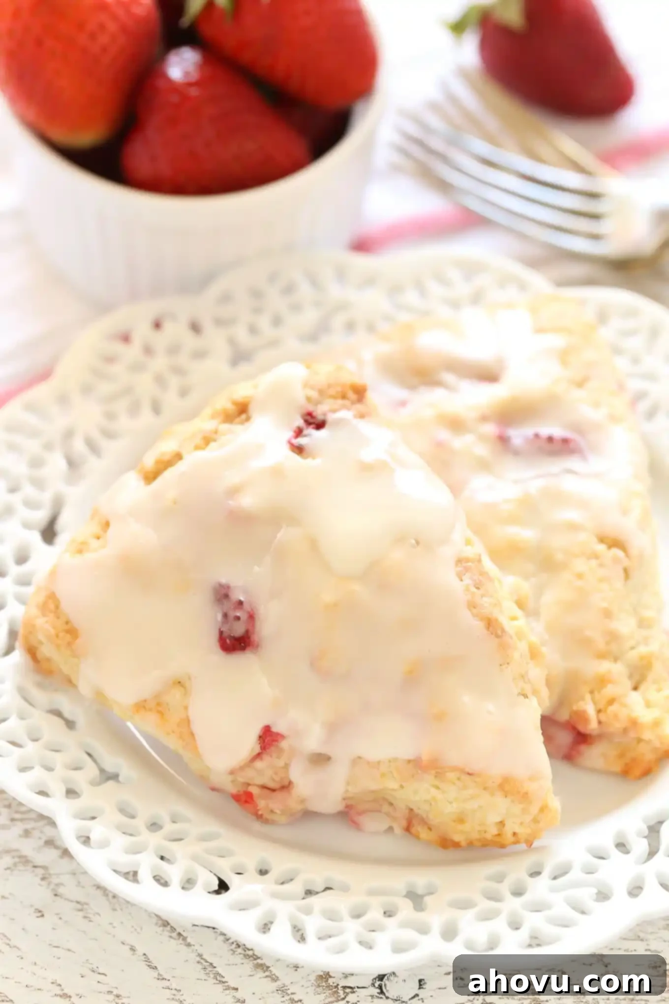 Homemade Strawberry Cream Scones 6 Two perfectly glazed strawberry scones elegantly presented on a white plate, accompanied by a small bowl of fresh berries and two forks in the background.