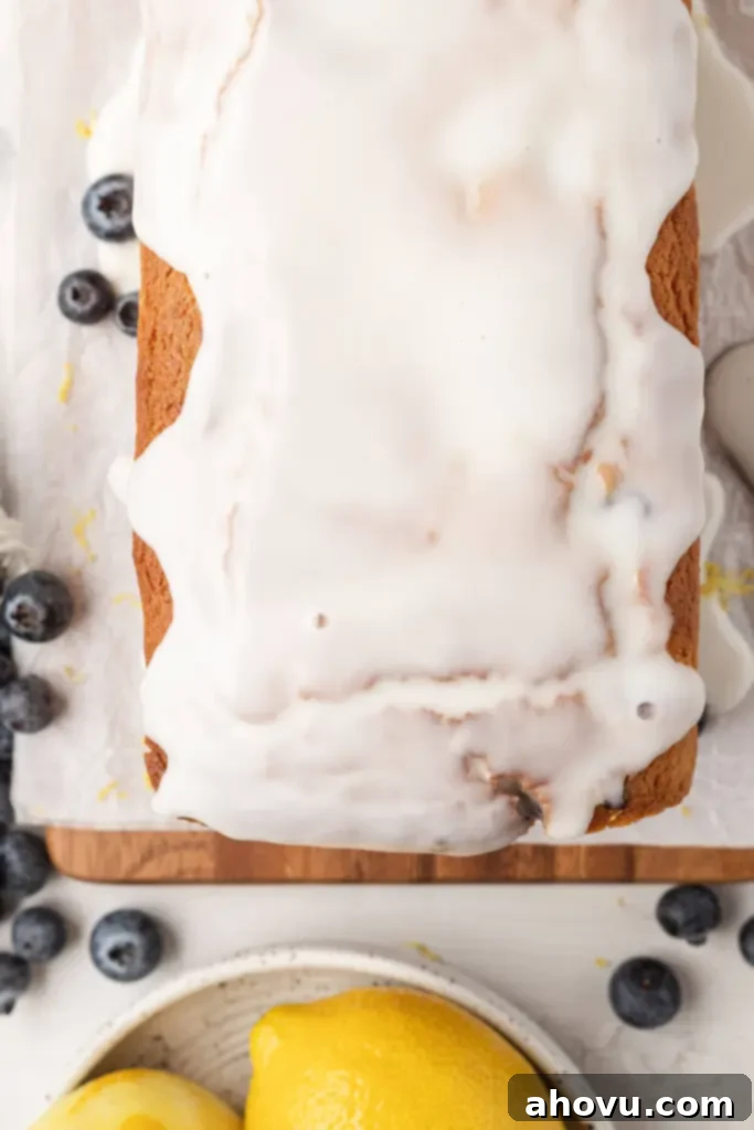 An overhead view of a freshly baked loaf of lemon blueberry bread with a shiny lemon icing, sitting on a wooden board surrounded by fresh blueberries.