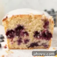 A straight on view of a loaf of lemon blueberry bread on a piece of parchment paper. The bread has been sliced to show the interior texture with scattered blueberries. A bowl of blueberries rests in the background.