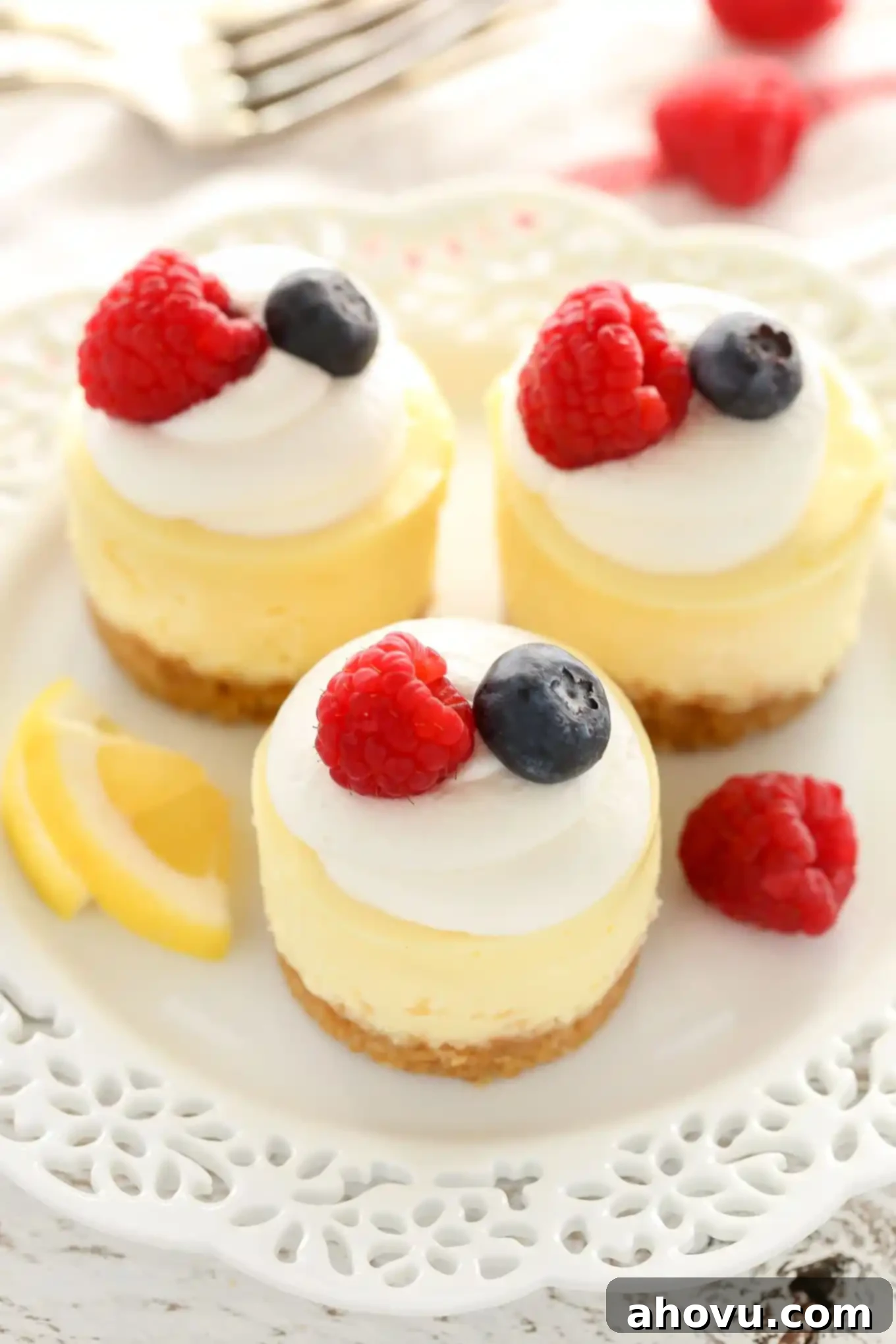 Overhead view of three mini lemon cheesecakes garnished with whipped cream and berries on a white plate. A fork rests in the background. 