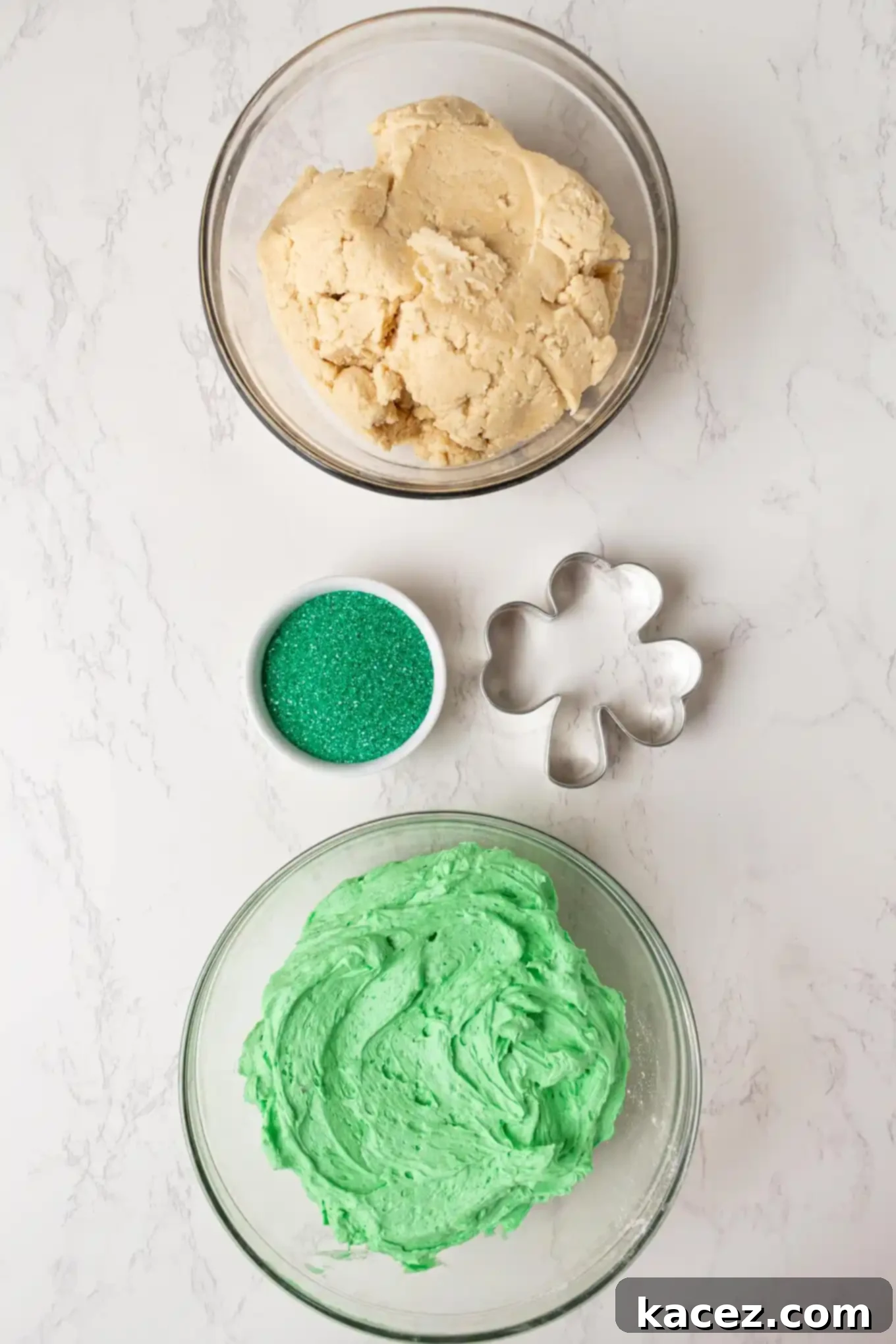Overhead shot of sugar cookie dough, vibrant green sanding sugar, smooth buttercream frosting, and a classic shamrock cookie cutter ready for St. Patrick's Day baking.